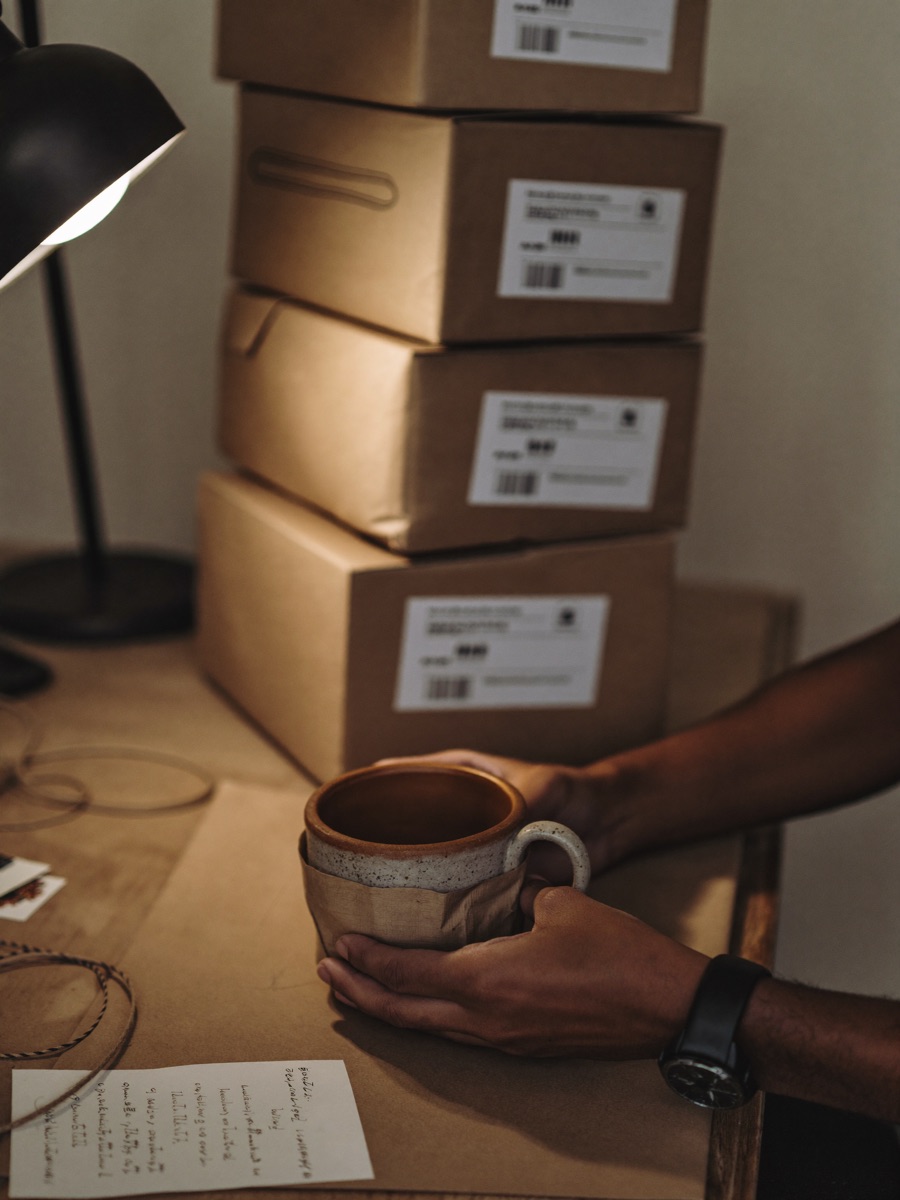 A maker's hands wrapping a handmade ceramic mug in kraft paper beside a stack of shipping boxes